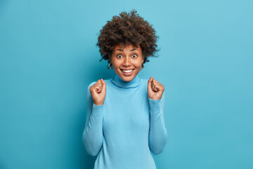 Happy dark skinned woman enjoys wonderful life and clenches fists with joy. Millennial girl...