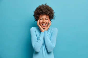 Fotobehang Young happy beautiful African American woman stands joyous with closed eyes, laughs as hears funny joke and touches cheeks. Cheerful positive millenial girl glad to spend free time with friends © Wayhome Studio