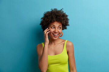 Pleasant looking happy woman with Afro hairstyle has nice telephone conversation and looks aside, wears casual vest. Ethnic girl has nice mood and smiles positively while talks via cell phone