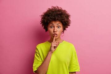 Surprised dark skinned woman makes silence gesture, keeps finger over lips and looks mysterious at...