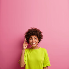Vertical shot of positive dark skinned woman with curly hair points above on blank space, dressed...