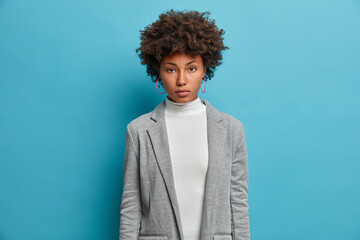 Fototapeta na wymiar Beautiful serious confident African American woman employee looks directly at camera with confident expression, wears formal clothes and poses against blue background. Female recommends good sale