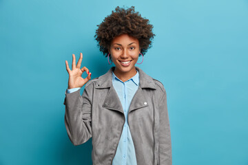 Confident self assured woman shows okay sign or all right gesture, recommends good product and smiles positively. Happy smiling lady in grey jacket gestures against blue wall. Body language concept