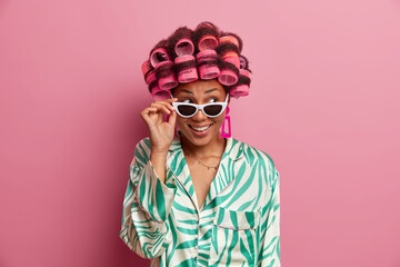 Horizontal shot of pretty young woman in sunglasses and silk dressing gown, makes hairstyle with hair rollers. Pleased housewife prepares for date or special event in life. Beauty time concept