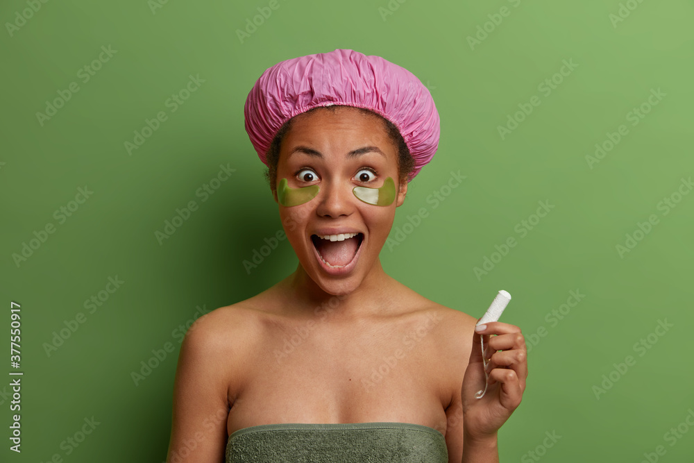 Wall mural Isolated shot of crazy surprised dark skinned female model with opened mouth holds tampon for protection during menses, enjoys beauty treatments after taking shower and poses against green background.