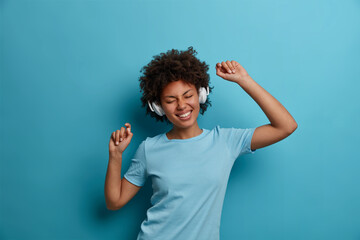 Carefree joyful young woman with Afro hair listens cool new favorite song in stereo headphones and dances carefree, closes eyes in pleasure. Cheerful female music lover wears casual blue t shirt