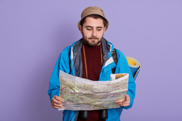Young bearded explorer man doubting and confused, holding map in hands and looking at it with pensive look, guy wearing jacket and hat, has blue mat behind back.