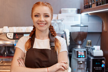 Fototapeta na wymiar Young smiling red haired barista standing at bar in coffee shop, keeps hands folded, looks at camera, wearing white casual t shirt and brown apron, standing near coffee machine.