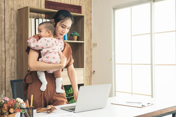 Asia mother stand and working online with a laptop holding her baby daughter at home office. Serious single mom with her child. Asian woman working from home, while in during Covid-19 crisis.