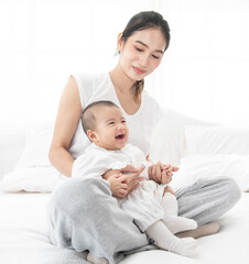 Asian mother and baby laugh together at home. Asia mom holdind infant and sitting on white bed in living room at house  having fun together.
