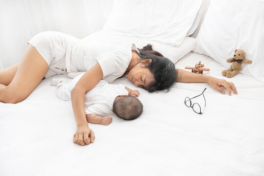 Tired Exhausted Mother Sleeping Sleep With Baby On White Bed. Overwhelmed New Asian Mom Feeling Sleep Deprived And Fatigued With Infant At Bedroom.