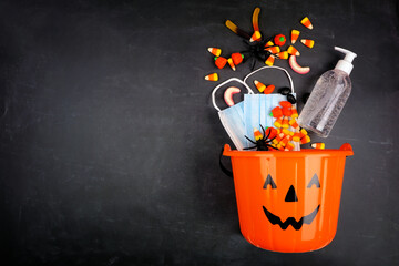 Halloween Jack o Lantern pail with spilling candy and COVID 19 prevention supplies. Top view over a black background.