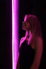 Club style portrait of girl in a black dress.   lit with violet light. Picture has dark  tone. Wall of neon has violet and white color,   outside next to night club, neonity city lights, minimalistic