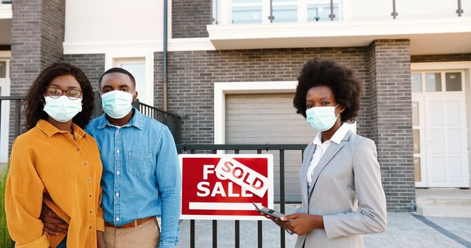 African American Young Happy Married Couple In Medical Masks Standing Outdoor At New House With Female Real-estate Agent Smiling To Camera. Sold Home. Pandemic Concept. Buying Dwelling.