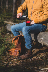 Ein Mann auf Wanderung macht eine Pause im Wald, während er auf einem Baumstamm sitzt und sich ein Heißgetränk aus seiner Thermoskanne in den Becher gießt.