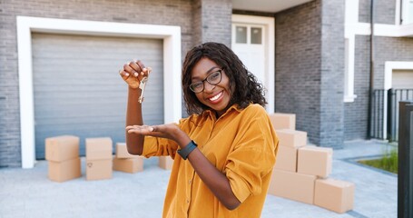 Portrait of cheerful happy African American woman in glasses smiling to camera and showing key to camera while moving in new home. Outdoor. Female demonstrating keys. Carton boxes on background.