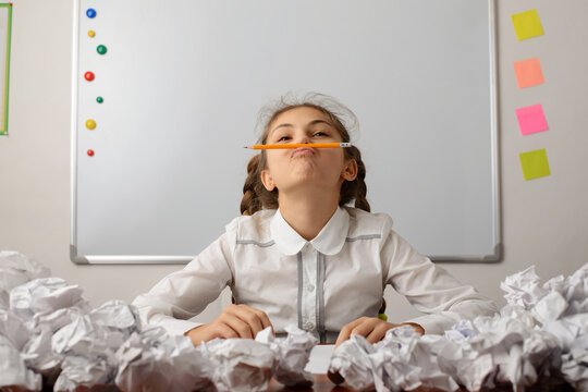Bored lazy young female student at the table with a lot of crumpled papers around her. Girl with two pigtails placing a pencil between her lips and nose. Concept of learning, boredom and laziness. - Powered by Adobe