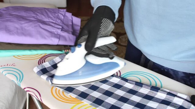 A man in gloves ironing the sleeve of a man's shirt. On the ironing board is a stack of men's shirts for ironing. Close-up shot.