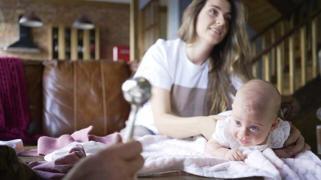 smiling brunette massages little bay girl back sitting on brown leather sofa and aged grandparent hand shakes blurred children rattle on foreground at home