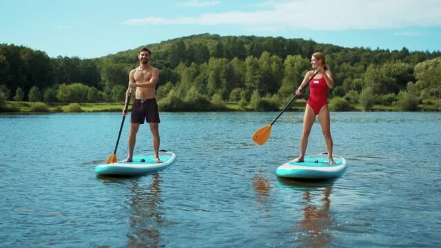At sunlight man and woman in a bathing suit standing on sup board and swims surfing on the lake with a paddle for swimming rowing sport surfer friends close up slow motion