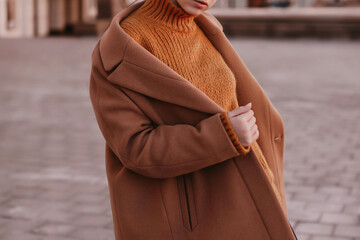 Young woman  with cropped face in an orange knitted sweater and brown coat outdoor portrait in daylight. Cozy autumn fashion style.  - obrazy, fototapety, plakaty