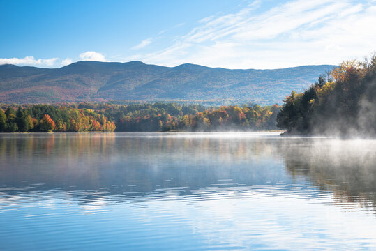 Morning fog drifting over the water of mountain lake with wooded shores at the peak of fall foliage. Waterbury, VT, USA.
