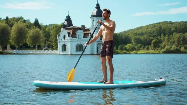 Attractive young man on sup paddle standing board at sunlight on lake summer water sport lifestyle travel boarding activity surfboard close up slow motion