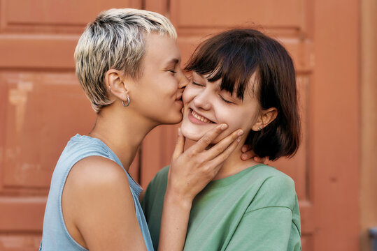 Pretty girl with short hair kissing her girlfriend outdoors. Young female couple smiling while standing in front of the door. LGBT, Sexual freedom concept