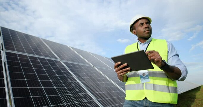 African-American worker engineer in uniform with white helmet walking in rows with solar panels making notes on tablet.