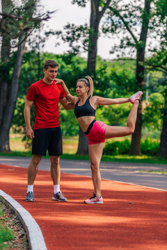 Young male athlete supporting his young female partner while she is stretching her legs after running on a race track.