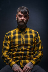 Tattooed man with a beard wearing checkered shirt is posing seated on a chair in a studio