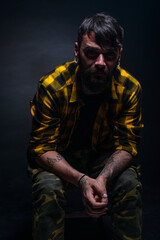 Obraz premium Tattooed man with a beard wearing checkered shirt is posing seated on a chair in a studio