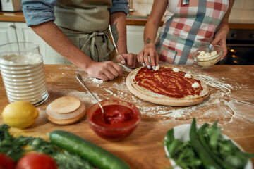 Add cheese. Cropped shot of couple making pizza together at home. Man in apron adding, applying tomato sauce on the dough while woman adding mozzarella cheese. Hobby, lifestyle