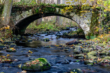 Fototapeta na wymiar A Water cascade in the creek flowing under an ancient stone bridge in the autumn forest.