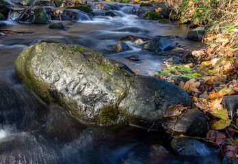 A water cascade with boulders in autumn creek with fallen leaves on a rocky shore. Water flows around the stones in the river.