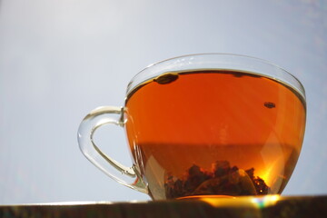 Transparent cup with black flower tea on the table. Background blue sky with sun.