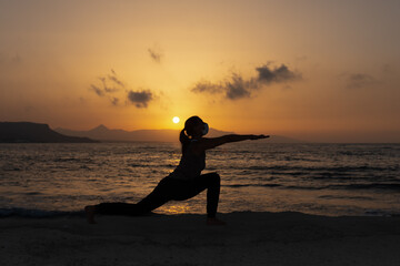 Silhouette, young woman with protective surgical face mask performs yoga stretching exercises at the beach at dusk during covid-19 coronavirus pandemic