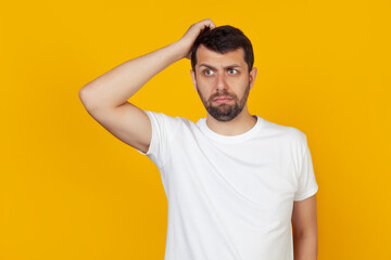 Fototapeta na wymiar Young man with a beard in a white t-shirt worried about a problem, with a hand on his head, nervous and anxious about the crisis, stands on an isolated yellow background