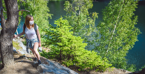 Young woman walking against the cliff with green trees and blue mountain lake wearing face protective mask for Covid 19 prevention. Traveler tourist with backpack. Corona virus and domestic tourism.