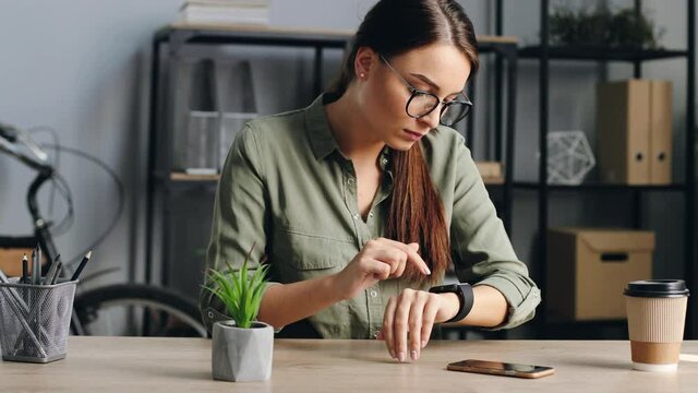 Close up of young beautiful woman with glasses using smartwatch at work. Attractive woman sitting in office configures electronic device.