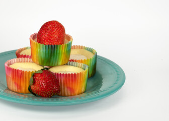 Five homemade cheesecake cupcakes in multicolored paper holders and fresh strawberries are served on a teal plate against a white background.