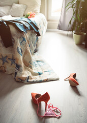 Red women's high-heeled shoes with pink panties on the white floor near the bed in a bright room