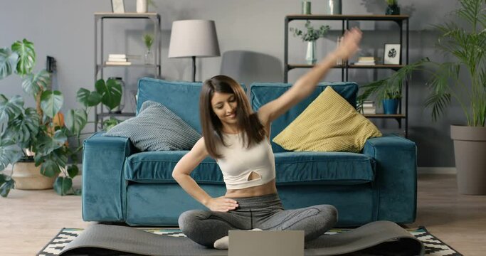 Pretty young Caucasian woman sitting on yoga mat on floor in front of the laptop computer at home. Attractive slim girl working out indoor with internet video course. Lesson with instructor online.