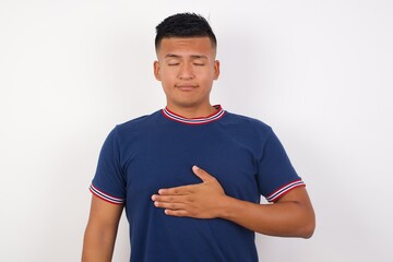 Young handsome hispanic man wearing casual t-shirt standing over white isolated background touches tummy, smiles gently, eating and satisfaction concept.