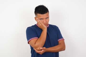 Very bored Young handsome hispanic man wearing casual t-shirt standing over white isolated background  holding hand on cheek while support it with another crossed hand, looking tired and sick,