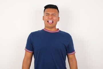 Young handsome hispanic man wearing casual t-shirt standing over white isolated background sticking tongue out happy with funny expression. Emotion concept.