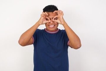 Young handsome hispanic man wearing casual t-shirt standing over white isolated background doing ok gesture like binoculars sticking tongue out, eyes looking through fingers. Crazy expression.