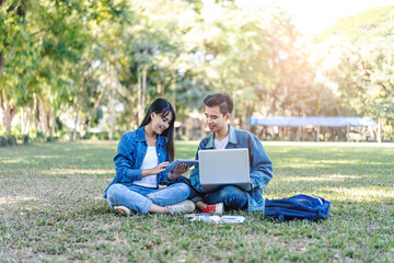 asian student couple studying outside on the grass, sitting down on the school campus using laptop computer, books, pencil and note book, studying hard under the trees nature in the evening sunlight