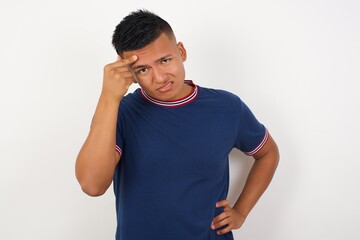Young handsome hispanic man wearing casual t-shirt standing over white isolated background pointing unhappy at pimple on forehead, blackhead  infection. Skincare concept.