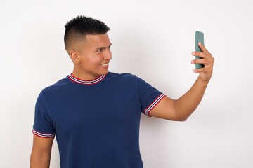 Charming Young handsome hispanic man wearing casual t-shirt standing over white isolated background, smiling and taking a selfie ready to post it on her social media.
